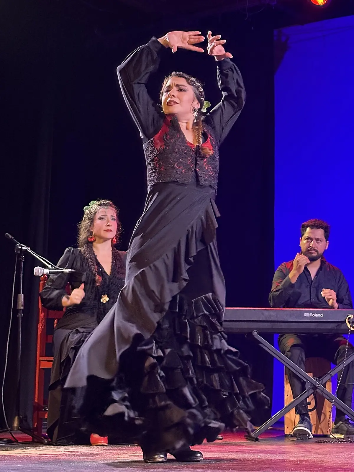 Flamenco tanečnice v černých volánových šatech zvedající paže na jevišti osvětleném modře, se zpěvákem sedícím vlevo a hráčem na klávesy vpravo