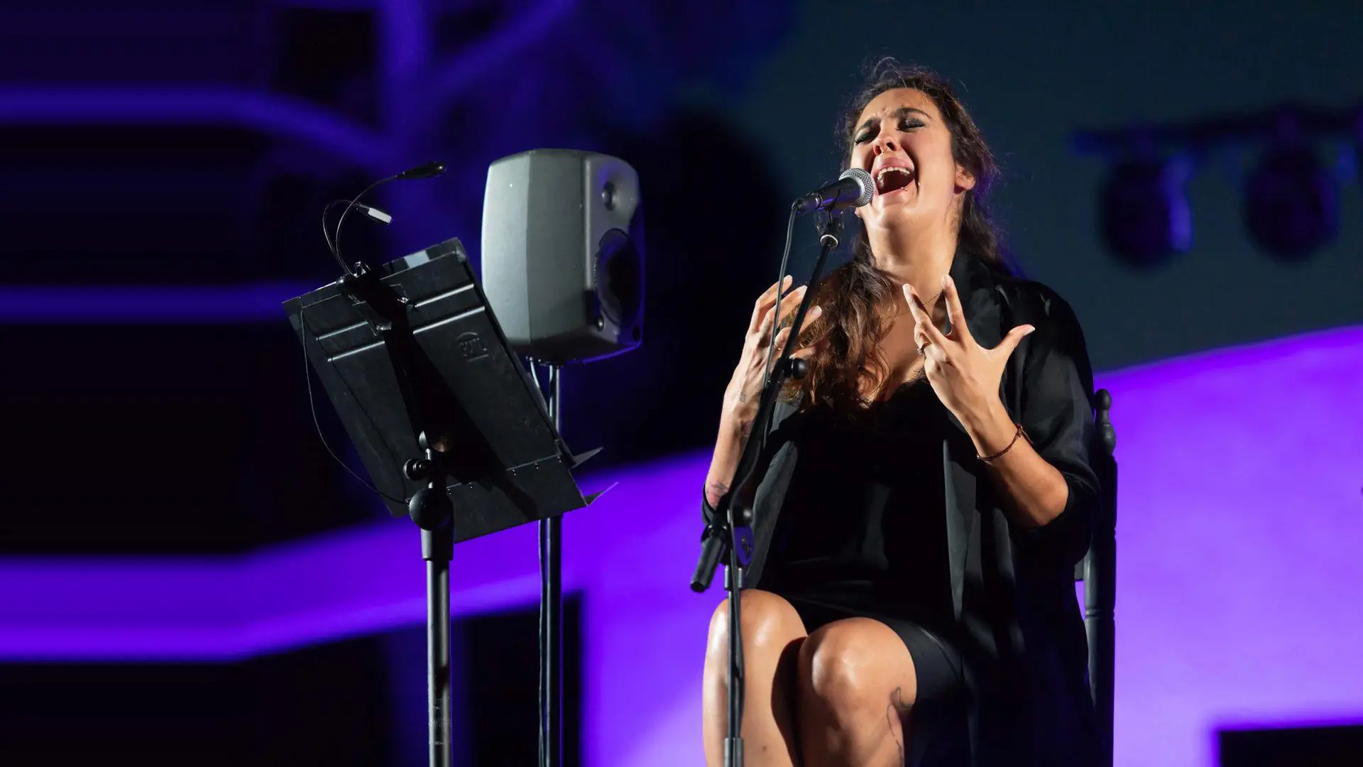 Flamenková zpěvačka sedící na jevišti se zdviženýma rukama, zpívá do mikrofonu ve fialovém světle