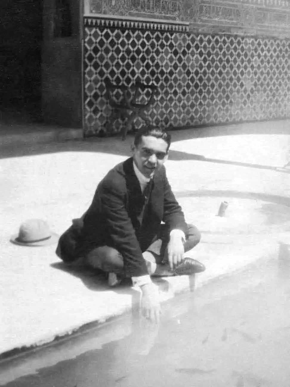Eine Schwarz-Weiß-Fotografie von Federico García Lorca, der am Rand eines Beckens in einem Innenhof der Alhambra sitzt, mit Fliesenwerk hinter ihm